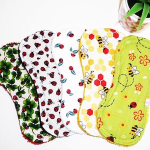 May include: Set of six reusable cloth menstrual pads with different patterns. The patterns include green four-leaf clovers, white ladybugs on a white background, green leaves on a white background, yellow and red honeycomb with bees, green flowers and bees on a green background, and red ladybugs on a green background.