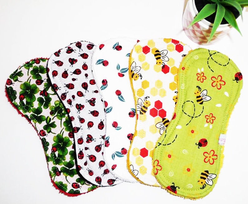 May include: Set of six reusable cloth menstrual pads with different patterns. The patterns include green four-leaf clovers, white ladybugs on a white background, green leaves on a white background, yellow and red honeycomb with bees, green flowers and bees on a green background, and red ladybugs on a green background.