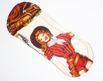 May include: A white fabric pad with a red and black checked design featuring a woman in a cowboy hat holding a gun. The woman is wearing a red and black checked shirt, a white scarf, and a brown belt. The fabric pad has a red stitched edge.