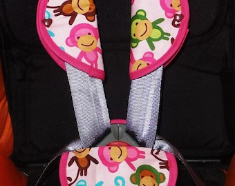 May include: A child car seat harness cover set. The covers feature a pink background with colorful monkey illustrations. The edges are trimmed with a bright pink fabric. The harness straps are gray.