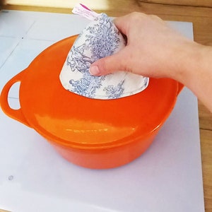 Può includere: Una pentola in ceramica arancione con un coperchio su un piano cottura bianco. Un copripentola in tessuto bianco con un motivo floreale è sopra il coperchio.