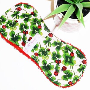 May include: A white cloth pad with a red border and a green four-leaf clover and ladybug print. The pad has a label that says "Mommy's" and "Fairy Magic".