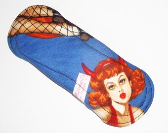 May include: A blue reusable menstrual pad with a retro devil girl illustration. The pad features a blue background with a red-haired woman wearing a red outfit and devil horns. The top of the pad has a fishnet pattern.