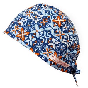 Puede incluir: Un gorro quirúrgico azul con un estampado floral en blanco, naranja y azul claro. El gorro tiene una corbata en la parte posterior y una pequeña etiqueta con texto.