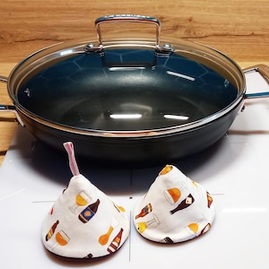 Può includere: Due presine in tessuto bianco con un motivo di bottiglie di birra e bicchieri. Le presine hanno la forma di un cono e sono appoggiate su una superficie bianca. Sullo sfondo c'è una pentola nera e argentata con un coperchio di vetro.