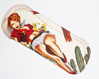 May include: A white fabric tube with a red, white and green design featuring a woman in a red and white outfit with a gun. The design also includes cacti and a wooden fence.