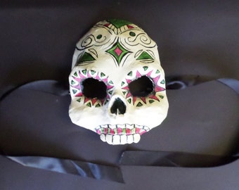 May include: A white skull mask with green and pink painted designs. The mask has eye holes and a black ribbon attached.