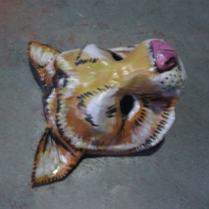 May include: A papier-m&acirc;ch&eacute; animal mask with a yellow, orange, and white striped pattern. The mask has a pink nose and black eyes.