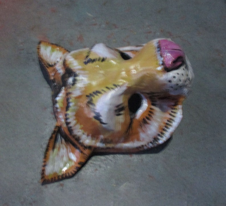 May include: A papier-m&acirc;ch&eacute; animal mask with a yellow, orange, and white striped pattern. The mask has a pink nose and black eyes.