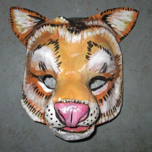 May include: A hand-painted tiger mask with orange, white, and black stripes. The mask has black eyes and a pink nose.