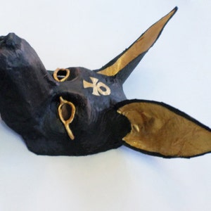 May include: A black Anubis mask with gold accents. The mask features large, pointed ears with gold interiors, gold eye details, and a gold ankh symbol on the forehead. The mask is made of a textured material.