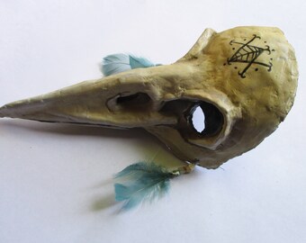 May include: A beige bird mask with a long beak and a black symbol on the side. The mask has two blue feathers attached to it.