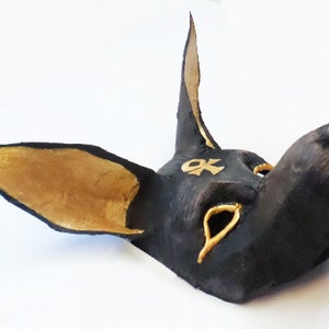May include: A black Anubis mask with gold accents. The mask features large ears, a long snout, and gold details around the eyes and on the forehead. The mask is likely handmade and has a textured appearance.