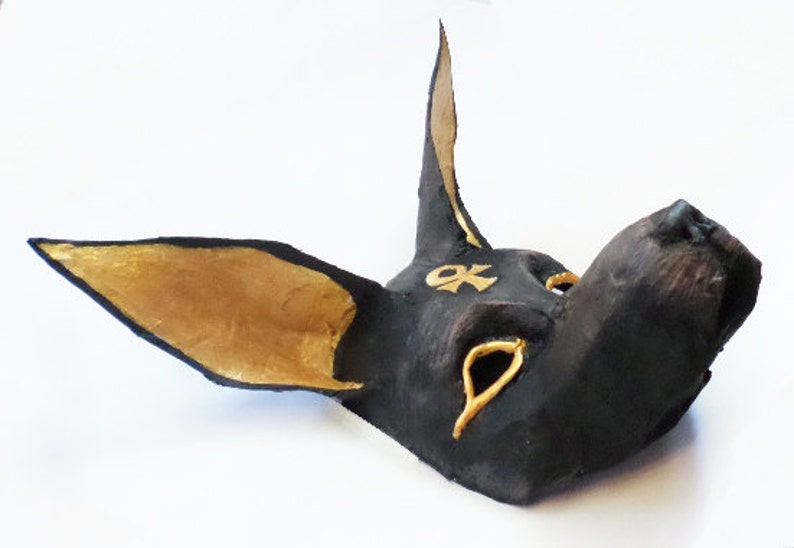 May include: A black Anubis mask with gold accents. The mask features large ears, a long snout, and gold details around the eyes and on the forehead. The mask is likely handmade and has a textured appearance.