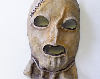 May include: A beige, paper mache mask with a stitched scar across the forehead, two eye holes, and a mouth hole. The mask has a textured surface and a frayed edge.