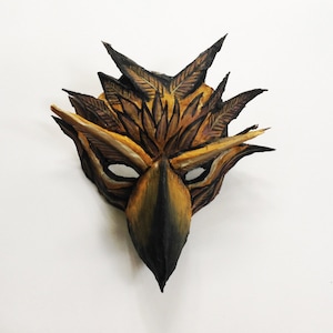 Könnte beinhalten: Eine braune und schwarze Vogelmaske mit einem Schnabel und Federn. Die Maske ist aus Papiermaché gefertigt und mit verschiedenen Farben bemalt.