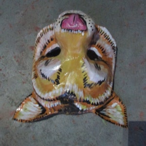 May include: A hand-painted tiger mask with orange, black, and white stripes. The mask has a pink tongue sticking out and black eyes.