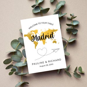 May include: White card with gold foil world map and the text "Welcome to your table Madrid Pauline & Richard August 28, 20XX". A paper airplane and a heart are drawn on the card.