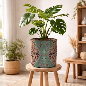 May include: A cylindrical fabric plant pot cover featuring a detailed geometric pattern in shades of teal, tan, and dark blue. The cover is placed on a light-coloured wooden stool and contains a green Monstera plant with large, split leaves.