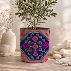 Könnte beinhalten: Ein zylindrischer Pflanzentopf mit einer lebendigen gewebten Textilaußenseite, die ein geometrisches Muster in tiefem Blau, leuchtendem Pink und Grün aufweist. Der Topf enthält einen kleinen Olivenbaum mit schmalen grünen Blättern und dünnen Ästen.