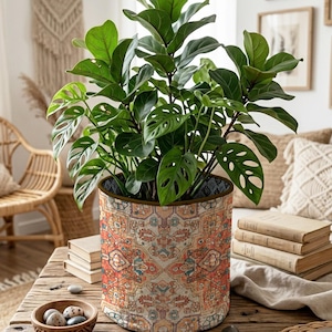 Puede incluir: Una funda de tela para maceta de colores vibrantes con un patrón geométrico simétrico y detallado en tonos naranja, azul y crema. La funda cilíndrica contiene una exuberante planta verde de hojas grandes tipo ficus lyrata.