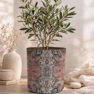Könnte beinhalten: Ein zylindrischer Pflanztopf mit einem detaillierten, sich wiederholenden Muster in Altrosa, Petrol und Creme. Das Design ahmt ein gewebtes Textil mit komplexen Blumen- und geometrischen Motiven nach. Ein kleiner Olivenbaum wächst aus dem Topf.