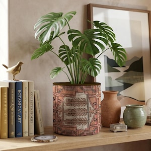 May include: A cylindrical fabric plant pot cover featuring a faded terracotta, beige, and dark grey geometric pattern. The pot holds a green monstera plant with large, split leaves. It is displayed alongside several books, a bird figurine, and vases.