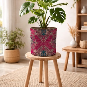 May include: A cylindrical fabric plant pot cover featuring a vibrant magenta, teal, and cream intricate floral pattern. The pot holds a green Monstera plant with large, split leaves. The cover has a brown rim and sits on a light wood stool.
