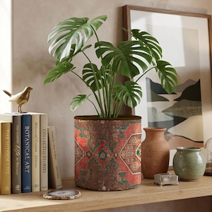 Op de afbeelding: Een cilindervormige plantenpot met een levendig geometrisch patroon in rood, bruin en blauwgroen, dat doet denken aan een geweven tapijt. De pot bevat een groene monstera-plant met grote, ingesneden bladeren en gedetailleerde symmetrische motieven.