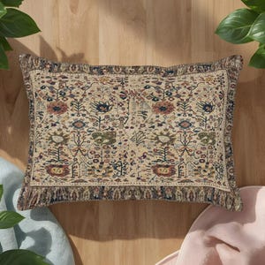 May include: A rectangular throw pillow featuring a beige base with a multi-colored floral and botanical pattern in shades of red, blue, green, and yellow. The pillow has a dark, intricate border and a textured fabric finish with a fringed edge.