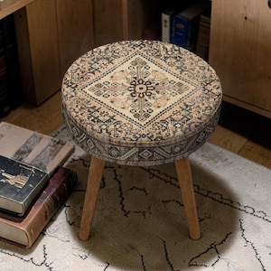 Può includere: Uno sgabello rotondo con un piano fantasia nei toni del beige, nero e marrone. Lo sgabello ha tre gambe in legno e un design decorativo che ricorda un tappeto vintage. È un elemento d'arredo decorativo.
