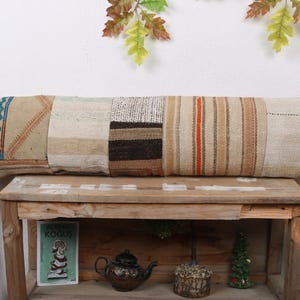 Peut inclure: Un long coussin rectangulaire avec un motif patchwork. Le coussin présente diverses teintes neutres, notamment beige, beige clair et marron, avec un mélange de textures et de motifs. Le coussin repose sur une table en bois.