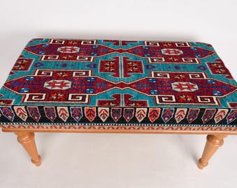 Könnte beinhalten: Ein Hocker aus Holz mit einer bunten, gemusterten Stoffoberfläche. Der Stoff zeigt ein geometrisches Muster in Rot-, Blau- und Weißtönen.