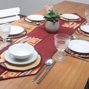Puede incluir: Un camino de mesa rojo y beige con patrones geométricos, colocado sobre una mesa de madera con platos blancos, cubiertos y vasos. El camino de mesa está decorado con una pequeña planta suculenta en maceta.