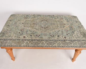 May include: A wooden bench with a light blue and cream patterned fabric top. The fabric has a floral design and a border around the edge. The bench has four legs.