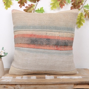 Pode incluir: Uma almofada quadrada bege com listras horizontais em tons de azul, laranja e cinza. A almofada é feita de um tecido texturizado e repousa sobre uma superfície de madeira. Folhas decorativas e um pequeno vaso com flores ao fundo.