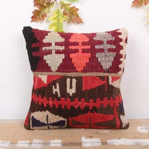 May include: A rectangular decorative pillow with a geometric pattern in shades of red, brown, black, and beige. The pillow is made of woven fabric and has a textured appearance. The pillow is placed on a wooden surface.