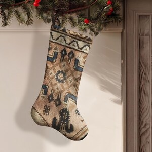 May include: A Christmas stocking featuring a tan, brown, and blue geometric pattern reminiscent of a woven rug. The stocking has a dark hanging loop at the top and displays intricate, symmetrical tribal-inspired motifs across its entire surface.