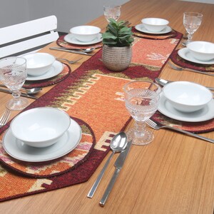 Puede incluir: Una mesa puesta con un camino de mesa estampado en rojo, naranja y verde. El camino de mesa tiene un diseño geométrico. Hay platos, cuencos y vasos blancos sobre la mesa.