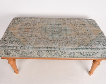 May include: A wooden ottoman with a light blue and beige patterned rug top. The ottoman has four turned legs.