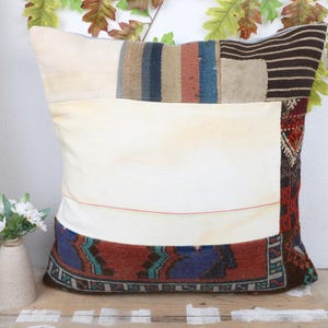 Puede incluir: Cojín decorativo cuadrado con diseño patchwork. El cojín presenta un centro color crema, acentuado con secciones de tela a rayas en azul, marrón y rojo. El borde inferior tiene un borde estampado en rojo, azul y verde.