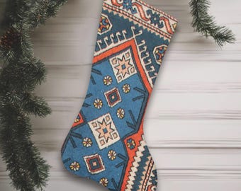 May include: A festive Christmas stocking with a blue, orange, and cream geometric pattern, suspended by a silver string. The stocking features a traditional rug design. A Christmas tree branch, pinecone, and red bauble are in the background.