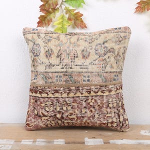 May include: A square decorative pillow with a vintage rug design. The top section is beige with floral patterns, the bottom is burgundy with a similar design, and a brown band separates the two. The pillow is placed on a wooden surface.