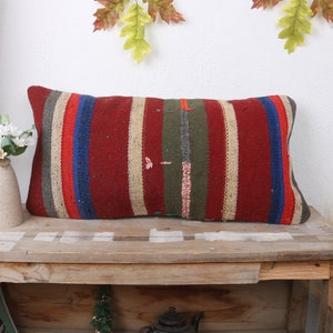 May include: A long rectangular pillow with a striped pattern in shades of red, beige, blue, and green. The pillow is made of woven fabric and has a rustic, vintage look.