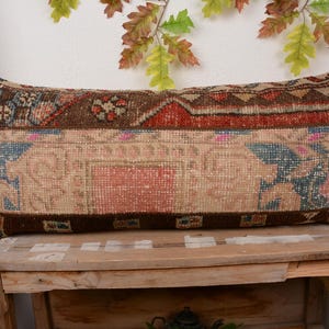 May include: Rectangular decorative pillow with a vintage rug design. The pillow features a mix of colors including brown, red, and blue. The pillow is placed on a wooden surface, with decorative leaves in the background.