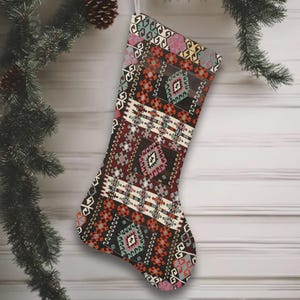 Puede incluir: Un calcetín navideño con un colorido patrón geométrico en tonos rojos, marrones, naranjas y turquesas. El calcetín cuelga de un lazo plateado sobre un fondo de madera blanca, con una rama de pino y adornos en el fondo.
