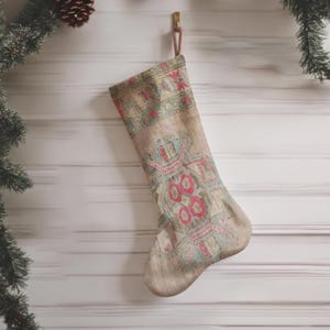 May include: A Christmas stocking made of light beige fabric featuring a woven geometric pattern in shades of muted red, teal, and cream. The stocking has a hanging loop at the top and displays a symmetrical, intricate design across its surface.