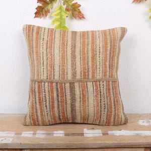 May include: Square decorative pillow with vertical stripes in shades of beige, brown, and orange. The pillow has a textured appearance and a horizontal band across the middle. The pillow is placed on a wooden surface.