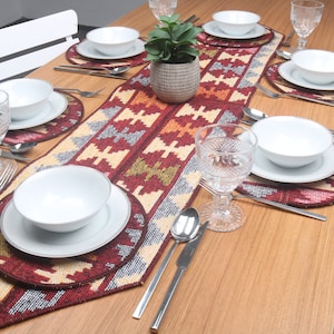 Könnte beinhalten: Ein rot-beige-grauer Tischläufer mit silbernen Akzenten. Der Läufer liegt auf einem Holztisch mit weißen Stühlen und verfügt über weiße Schalen und Teller, Besteck und Gläser.