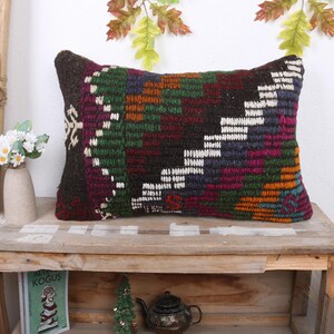 May include: Rectangular decorative pillow with a geometric pattern in shades of brown, green, purple, and orange. The pillow is placed on a wooden table, with a vase of white flowers and a small book visible in the background.